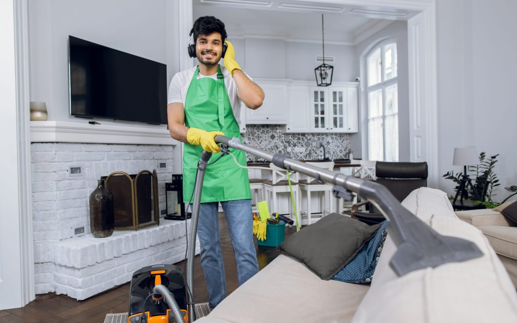 Smiling man cleaning service worker, cleaning sofa with vacuum and listening songs in wireless headphones. Indian guy wearing green apron and yellow rubber gloves.