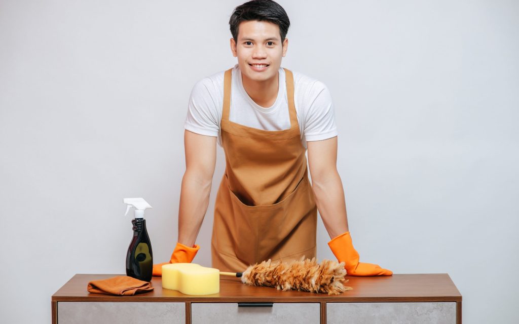 Happy Handsome man wearing apron and rubber gloves preparing to cleaning home, The feather duster, cleansing spray bottle, sponge and cloth wipper on cabinet with drawers, copy space